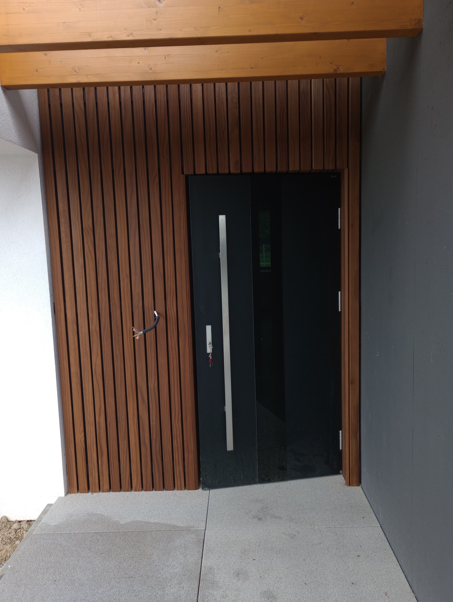 Nowoczesna elewacja domu z drewnianymi panelami pionowymi w odcieniach brązu, czarnymi drzwiami wejściowymi z długim, srebrnym uchwytem i surowym betonem, widoczne wystające przewody elektryczne po...