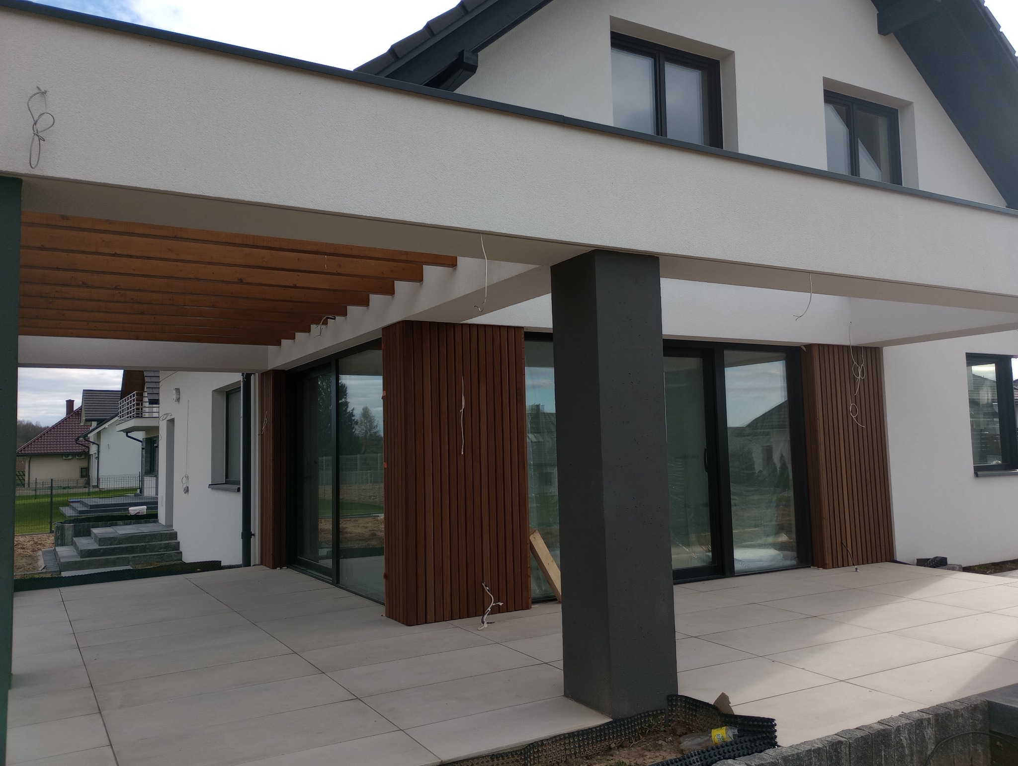 Nowoczesny dom z białymi ścianami i drewnianą pergolą nad tarasem wyłożonym jasnymi płytami, widoczne przesuwne okna i drewniane panele elewacyjne