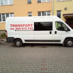 Biały bus z przyczepką zaparkowany na brukowanej nawierzchni, z czerwonym napisem 'TRANSPORT' i numerem telefonu na boku.