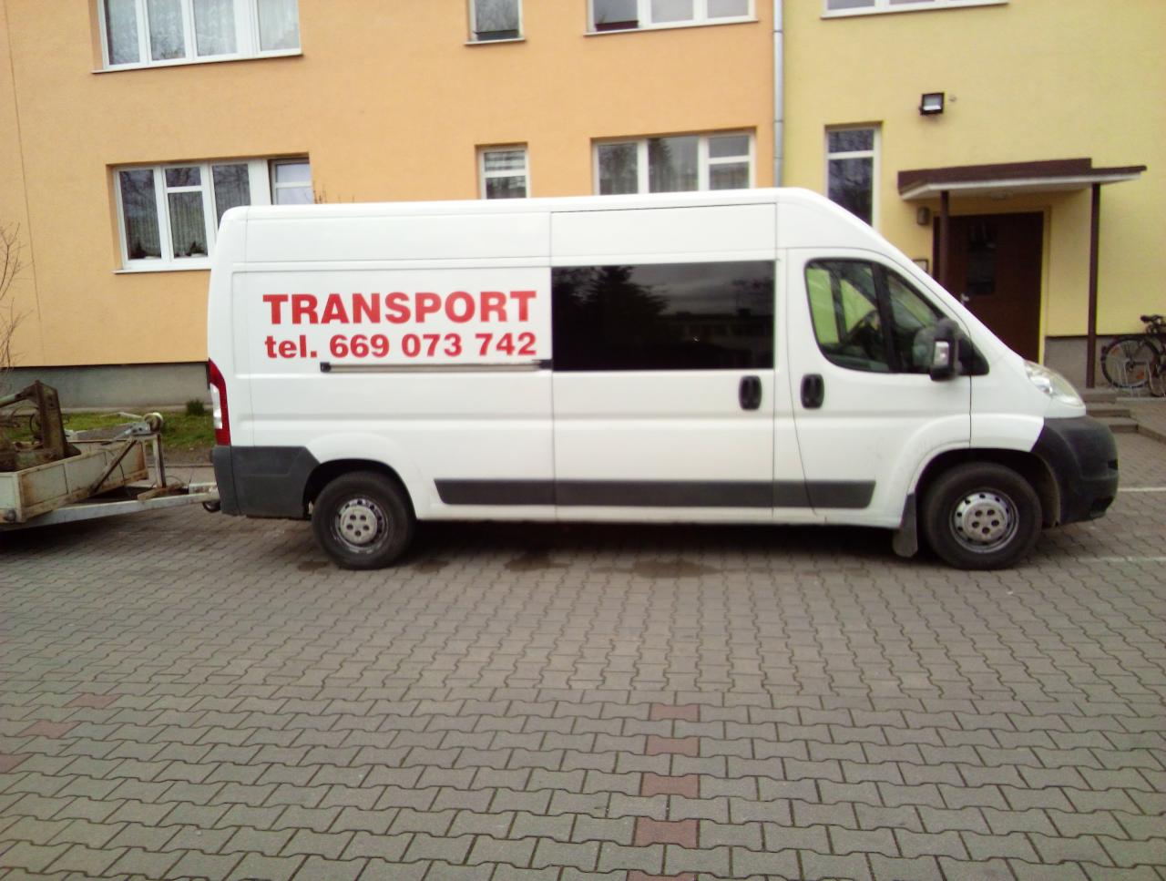 Biały bus z przyczepką zaparkowany na brukowanej nawierzchni, z czerwonym napisem 'TRANSPORT' i numerem telefonu na boku.