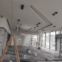 Malowanie sufitu w biurze, widoczne przygotowania: folia ochronna na podłodze, drabiny, sprzęt malarski i otwory na oświetlenie punktowe w suficie.