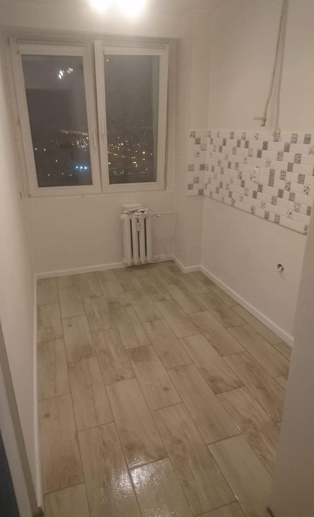 Wykończone pomieszczenie z beżową podłogą imitującą drewno, dekoracyjnymi płytkami na ścianie, białym grzejnikiem i oknem z widokiem na nocne miasto.