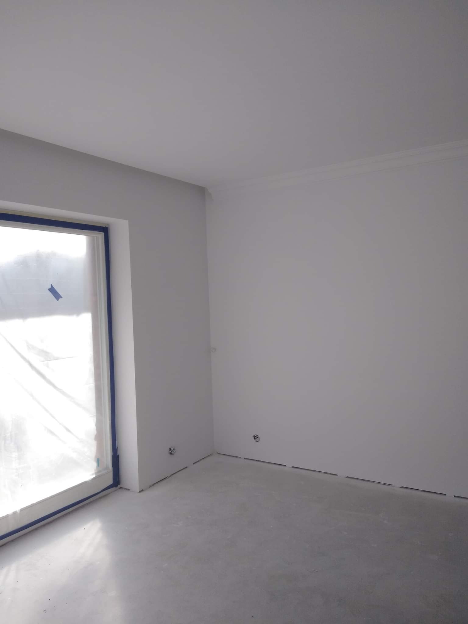 Pomalowane na biało wnętrze pokoju z zabezpieczonym folią oknem i niebieską taśmą malarską wzdłuż ramy okiennej i podłogi, widoczne gniazdka elektryczne.