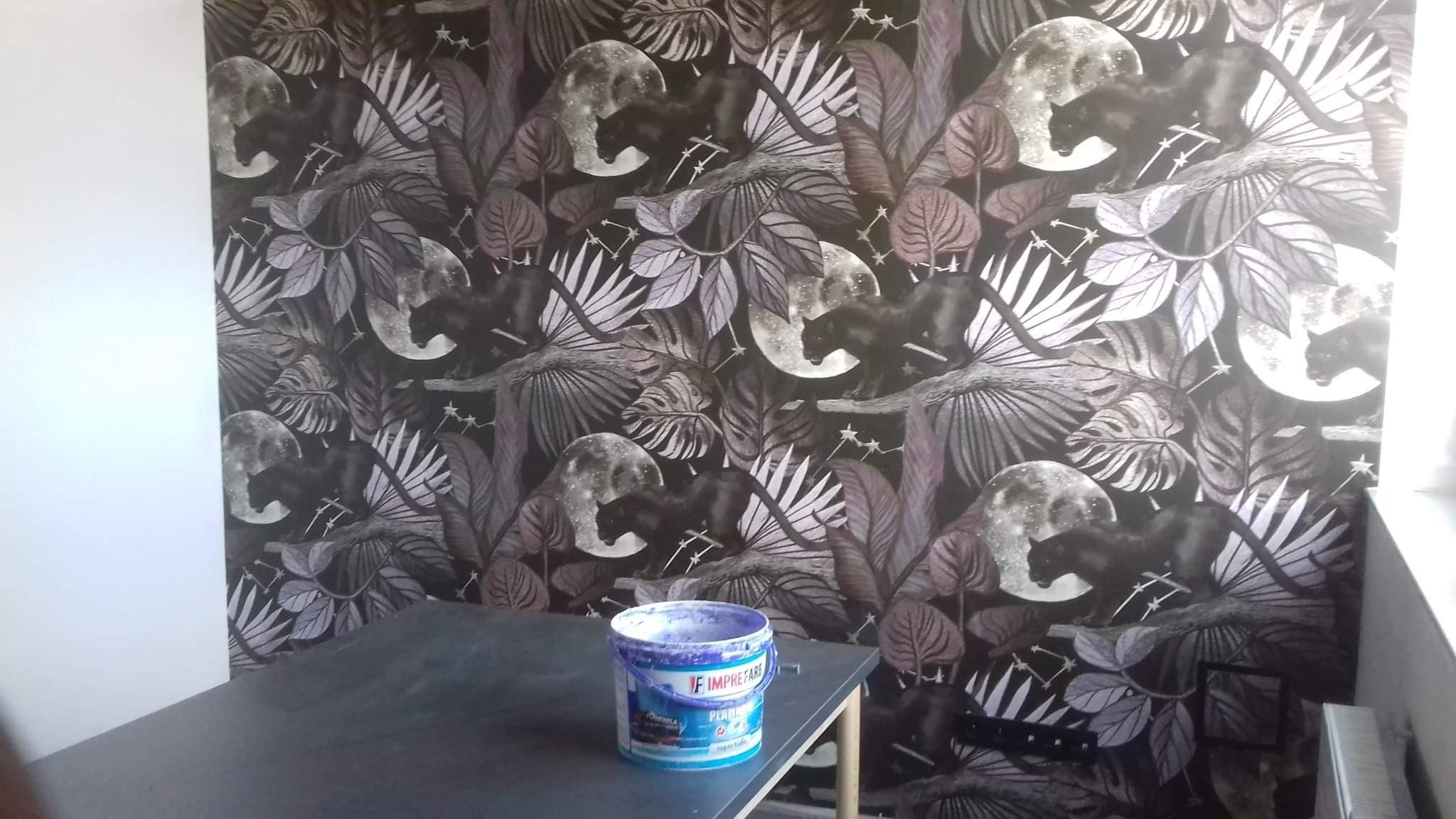 Ściana pokryta tapetą w motywy roślinne i panter, na pierwszym planie wiadro z farbą na stole.