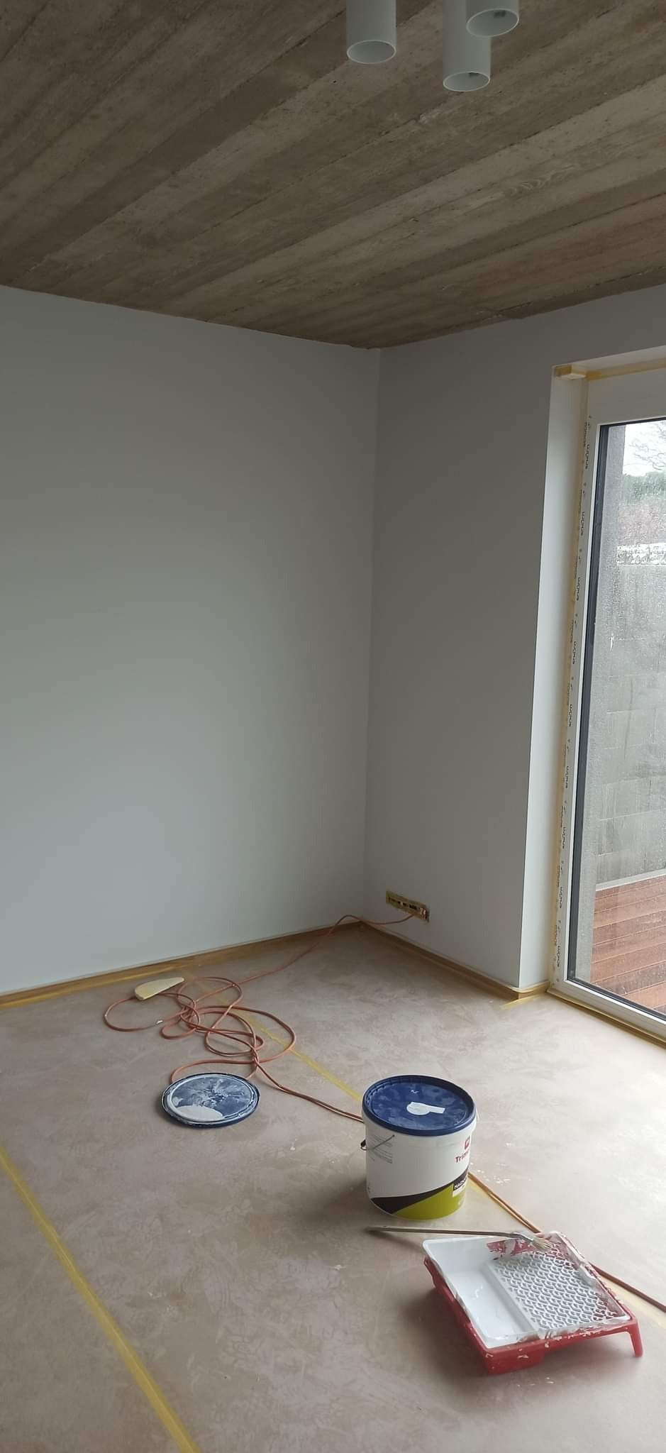 Pokój przygotowany do malowania, z taśmą malarską zabezpieczającą podłogę i okna, widoczne wiadro z farbą, kuweta malarska i kabel elektryczny.