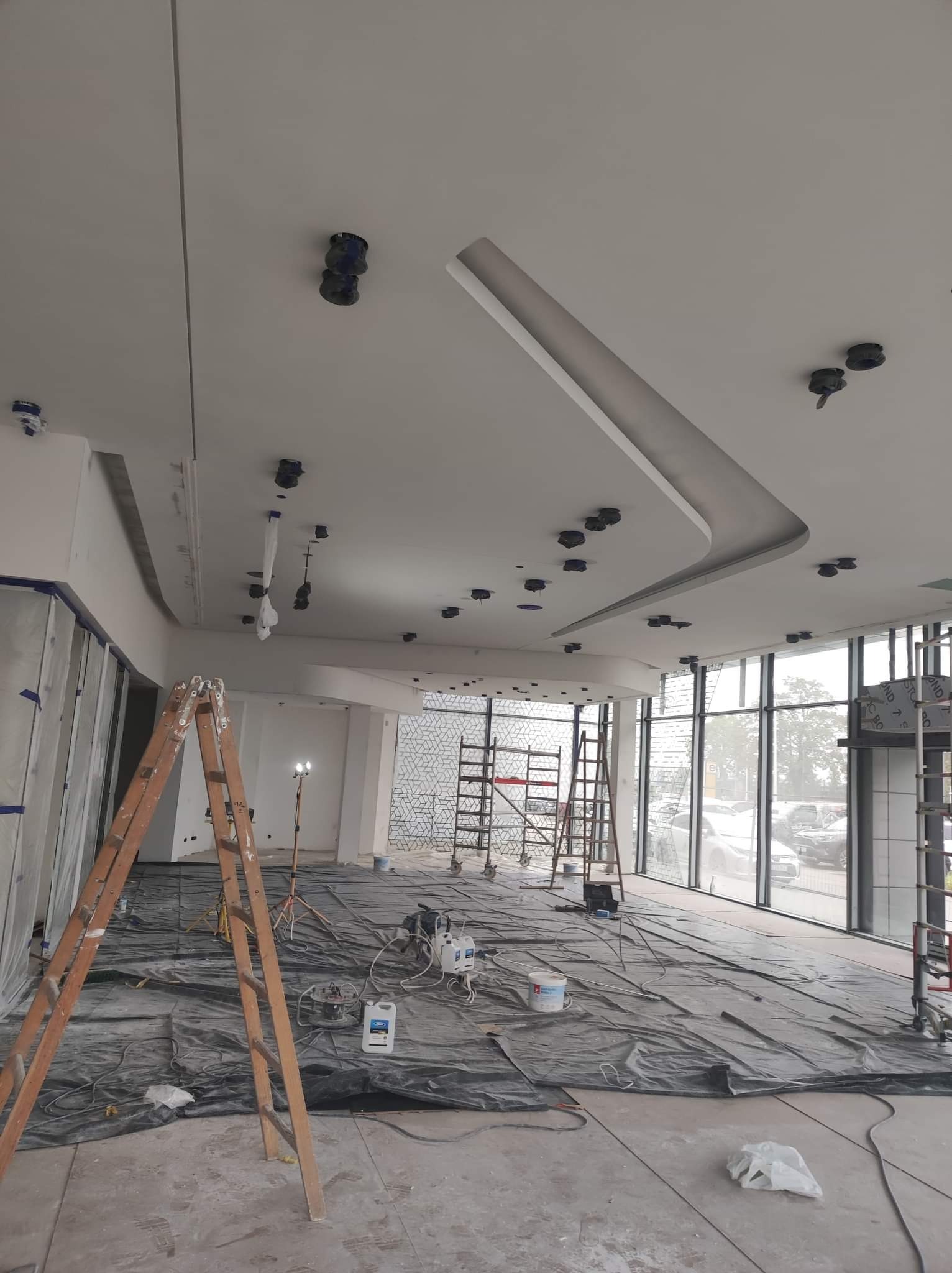 Malowanie sufitu w biurze, widoczne przygotowania: folia ochronna na podłodze, drabiny, sprzęt malarski i otwory na oświetlenie punktowe w suficie.