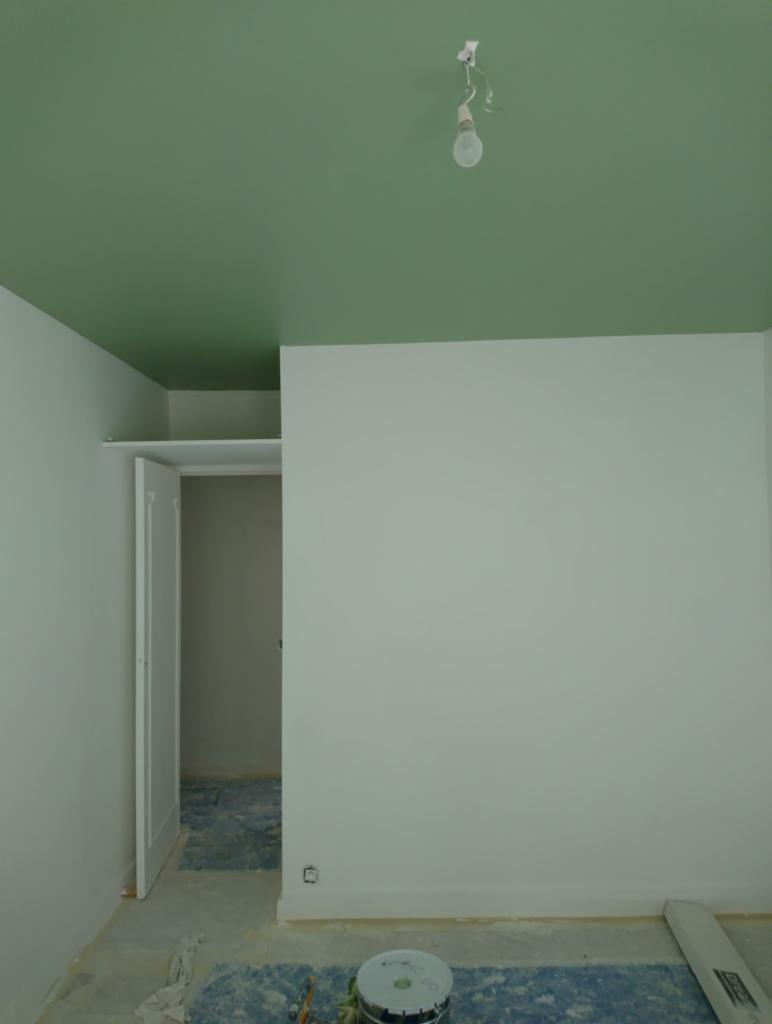 Pokój w trakcie odświeżania: zielony sufit, białe ściany, widoczne zabezpieczenia podłogi, otwarte drzwi do garderoby i puszka farby przy ścianie.