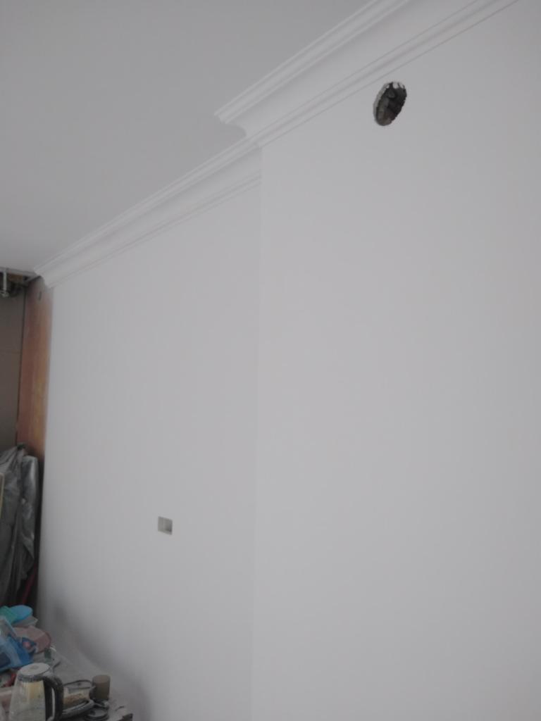 Świeżo pomalowane, białe ściany w rogu pomieszczenia z dekoracyjnym gzymsem pod sufitem i otworem na instalację elektryczną. Widoczne drobne niedoskonałości w wykończeniu.