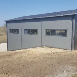 Szara hala z płyty warstwowej z trzema oknami, widok z zewnątrz na tle zaoranych pól i błękitnego nieba.