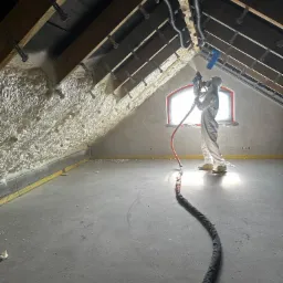 Pracownik w kombinezonie ochronnym natryskuje pianę izolacyjną na skośną ścianę poddasza, widoczne krokwie i okno w tle.