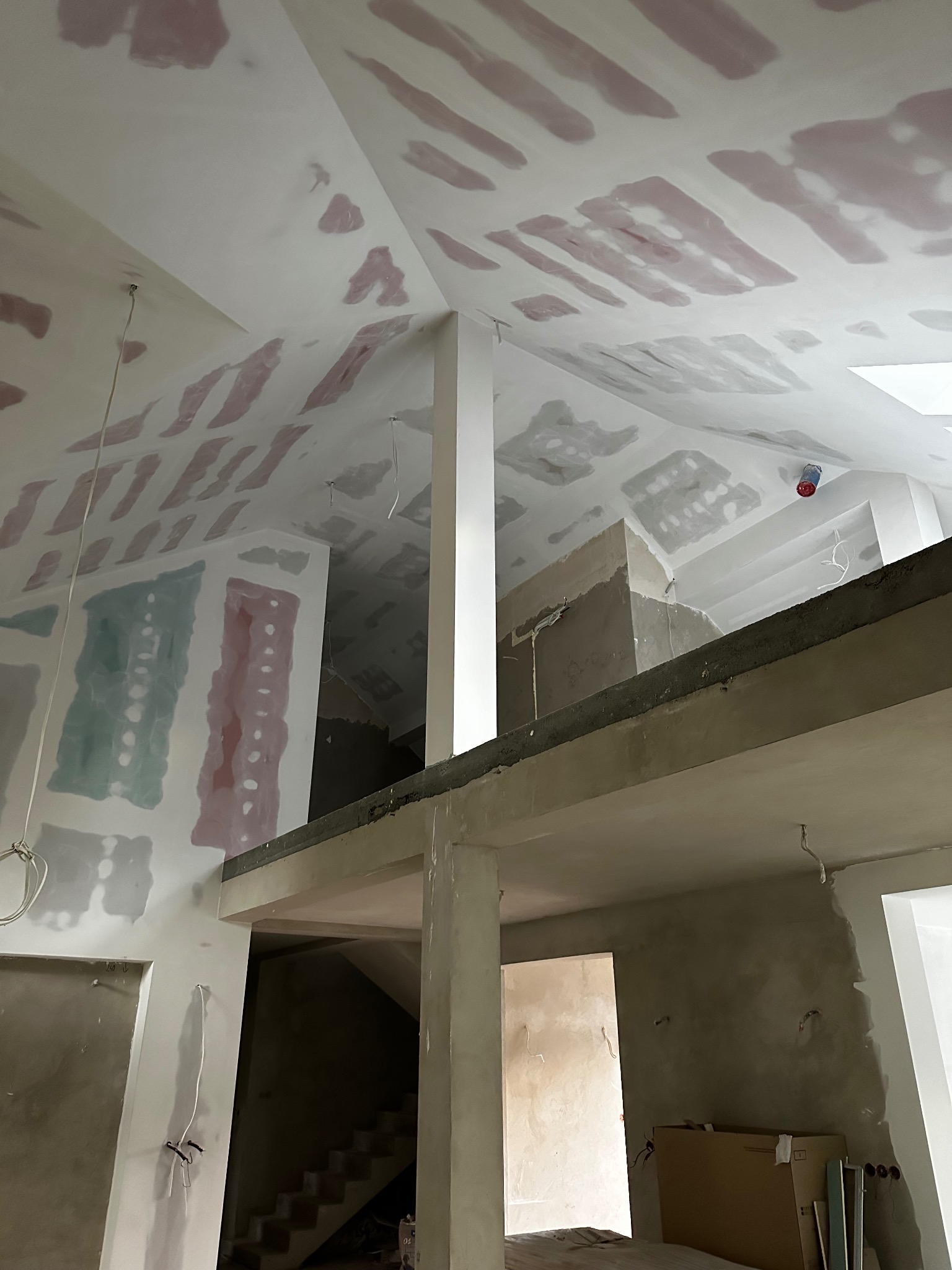 Wnętrze w trakcie remontu z widocznymi fragmentami ścian i sufitu pokrytymi wstępną warstwą masy szpachlowej w kolorze różowym i szarym, widok z dołu na antresolę z elementami instalacji elektrycznej.