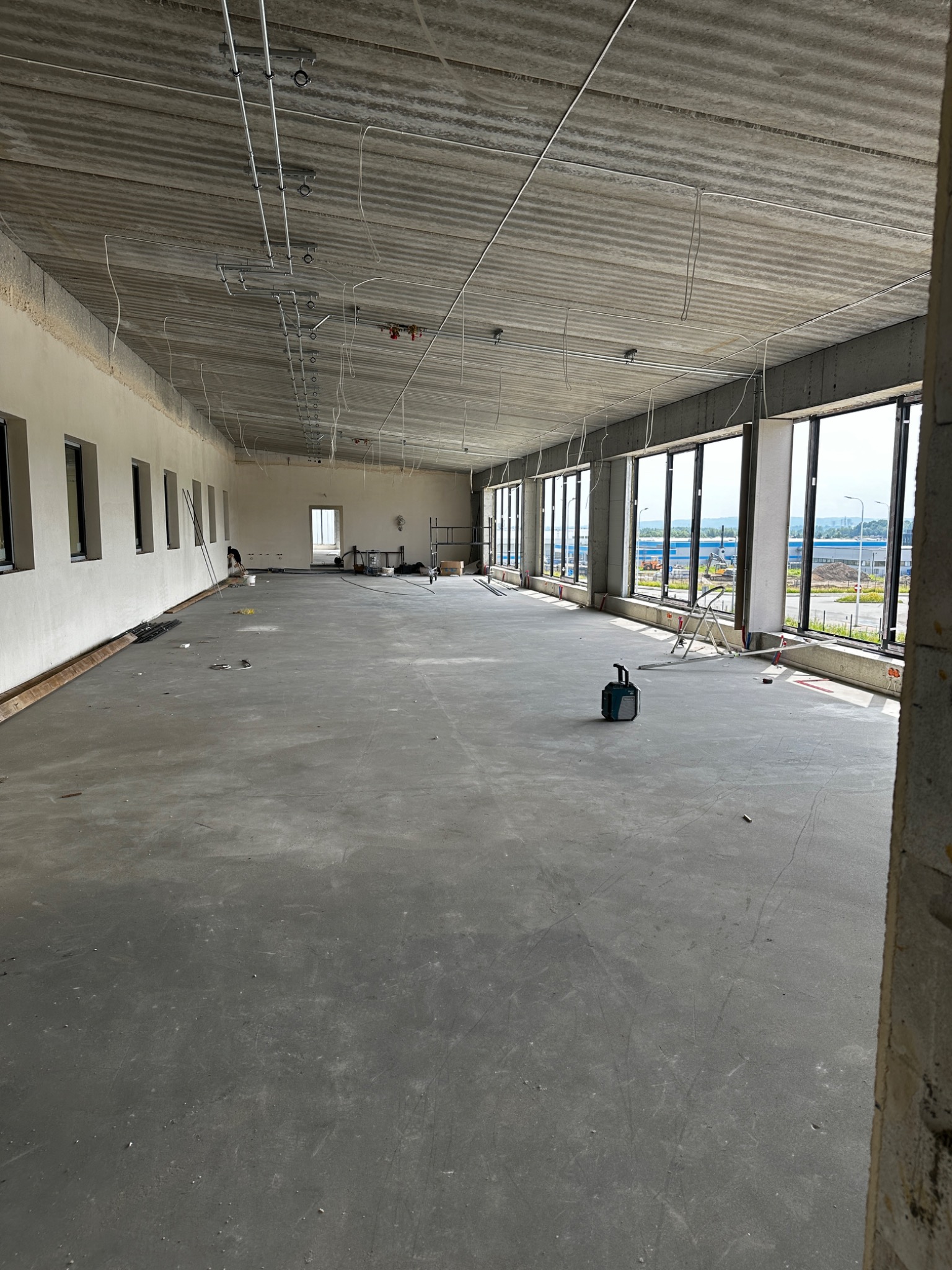 Wnętrze dużego, surowego pomieszczenia w trakcie prac wykończeniowych; widoczne okna, betonowa podłoga, instalacje pod sufitem i drabina; w tle budynki przemysłowe.