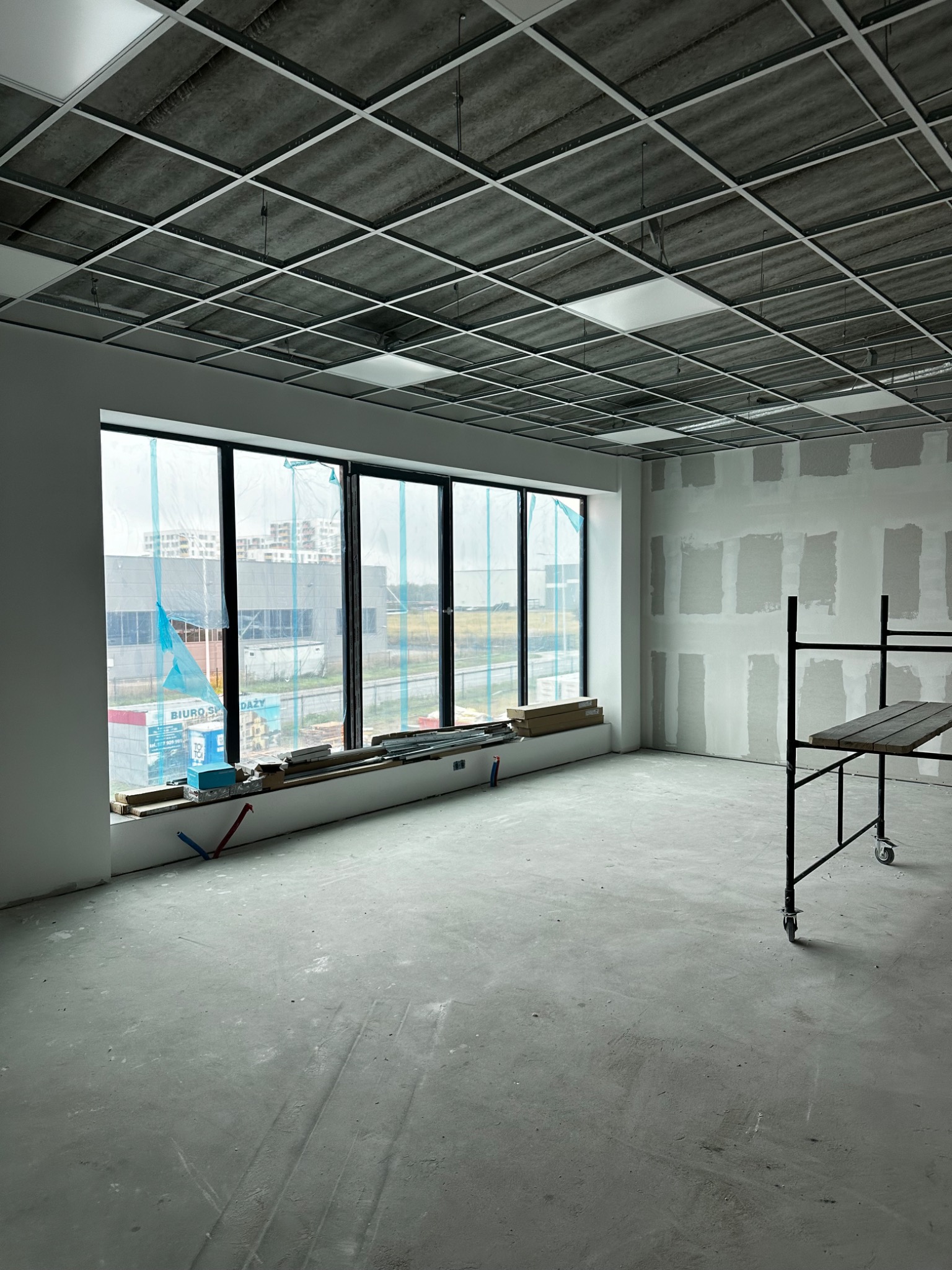 Wnętrze biurowe w trakcie prac wykończeniowych; widoczne szpachlowane ściany, sufit podwieszany, okno z widokiem na budynki oraz rusztowanie.