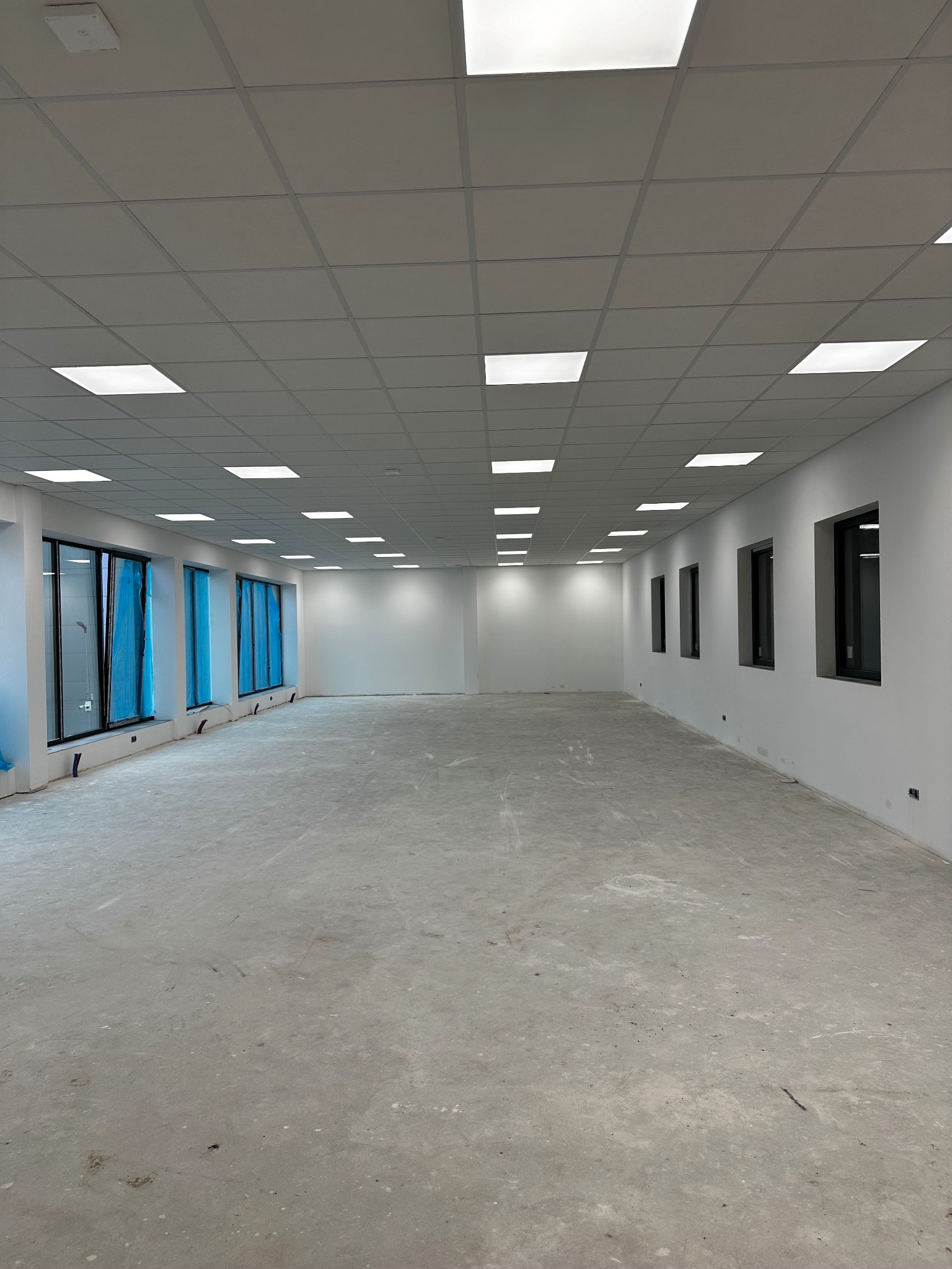 Wnętrze pustego, jasnego pomieszczenia biurowego z surowym betonem na podłodze i dużymi oknami zabezpieczonymi folią ochronną, przygotowanego do dalszych prac wykończeniowych.