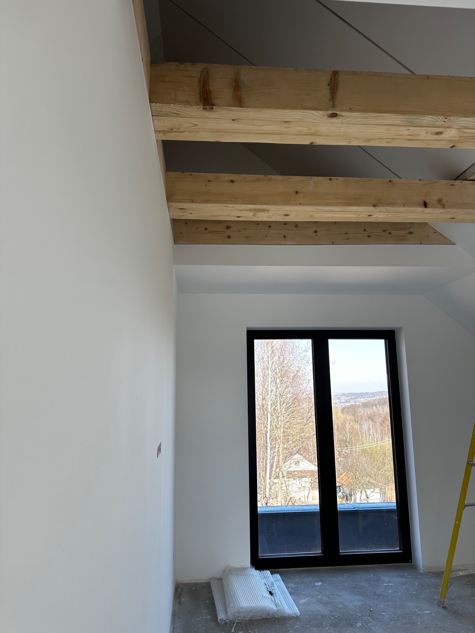 Poddasze ze świeżo pomalowanymi na biało ścianami, widoczne drewniane belki stropowe, czarne okno z widokiem na drzewa i fragment żółtej drabiny.