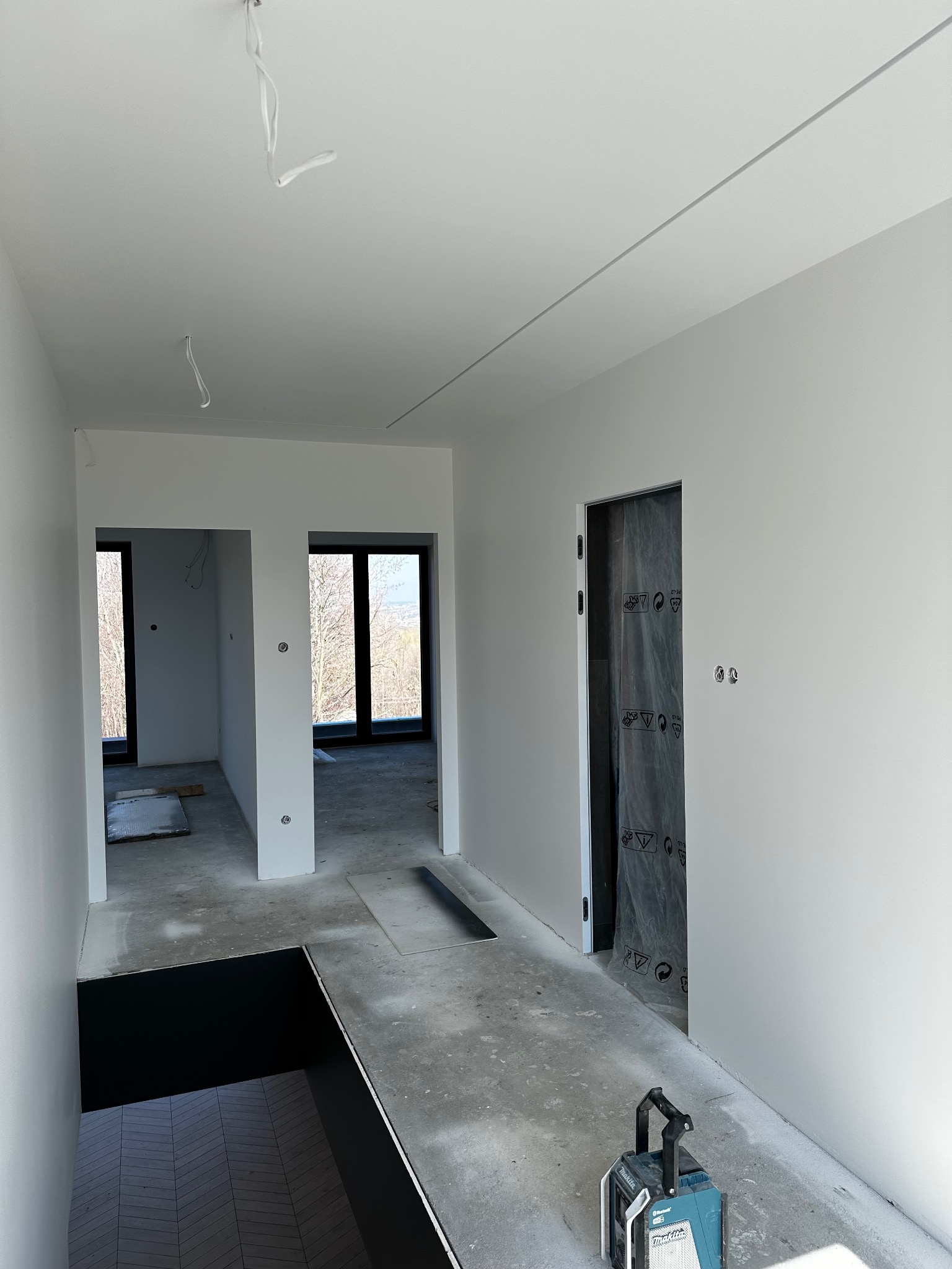Wnętrze w trakcie prac wykończeniowych: świeżo pomalowane, białe ściany, surowa podłoga, zabezpieczone drzwi folią, widoczny sprzęt budowlany i przygotowane otwory okienne.