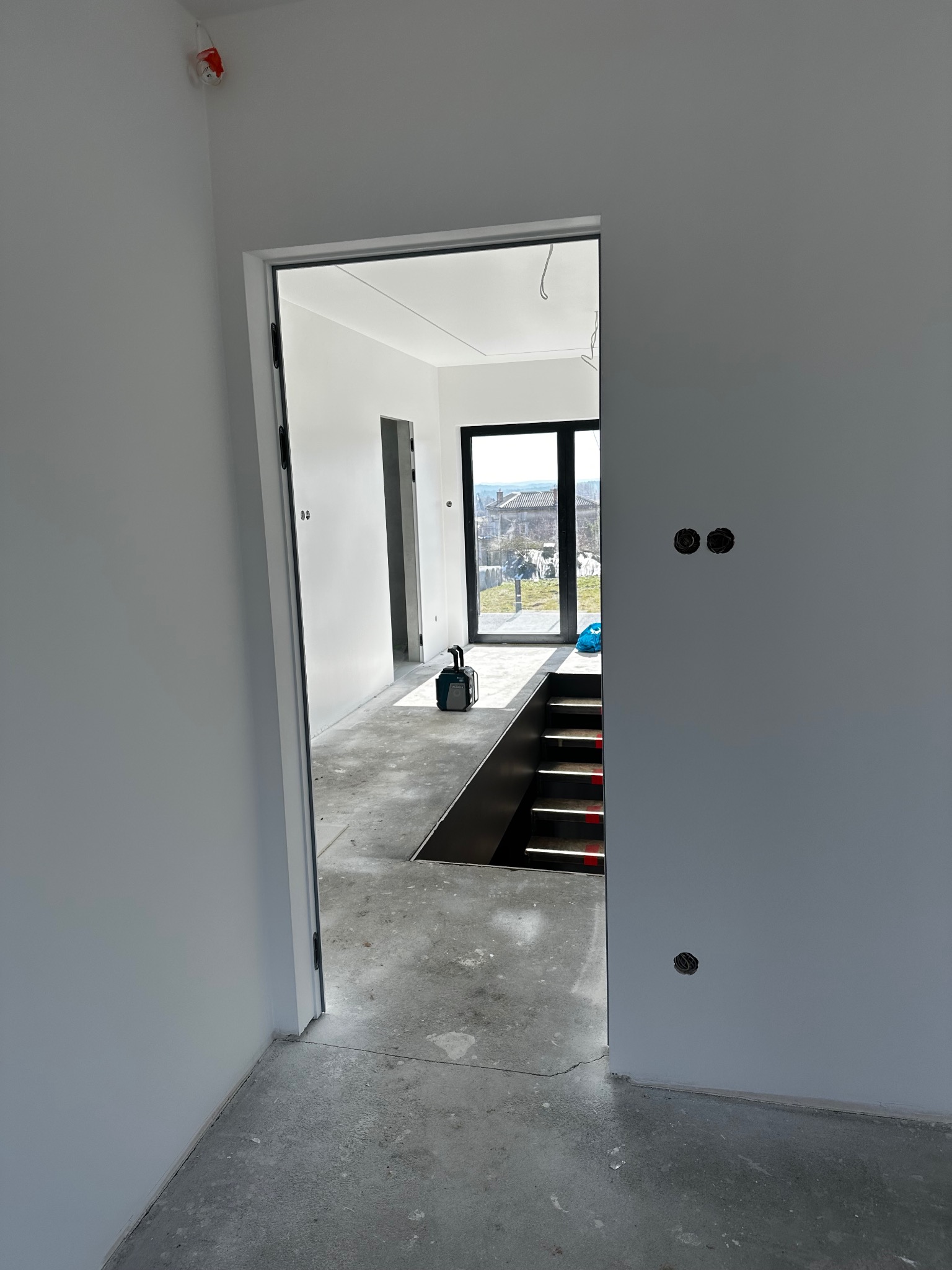 Wnętrze w trakcie remontu: surowe ściany pomalowane na biało, betonowa podłoga, widoczne schody z czarnymi barierkami i czerwonymi akcentami, otwory na gniazdka elektryczne i narzędzia budowlane...