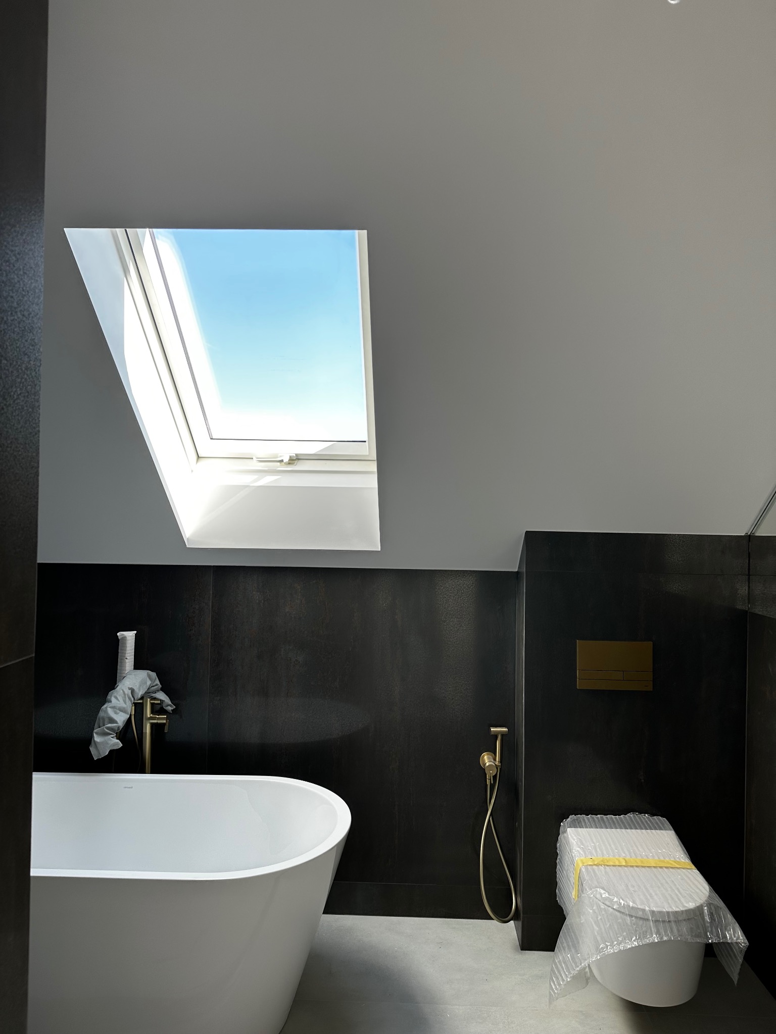 Nowoczesna łazienka w trakcie wykańczania, z wolnostojącą białą wanną, złotą armaturą, toaletą zabezpieczoną folią i oknem dachowym w skosie.