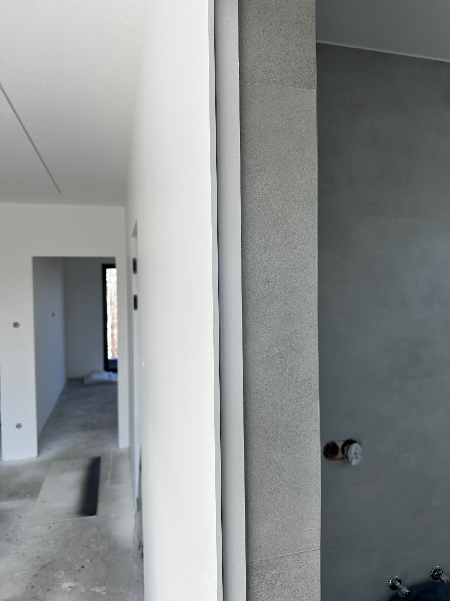 Fragment wykończonego wnętrza z białymi ścianami i szarą łazienką z widocznymi przyłączami hydraulicznymi, surowy beton na podłodze w tle.