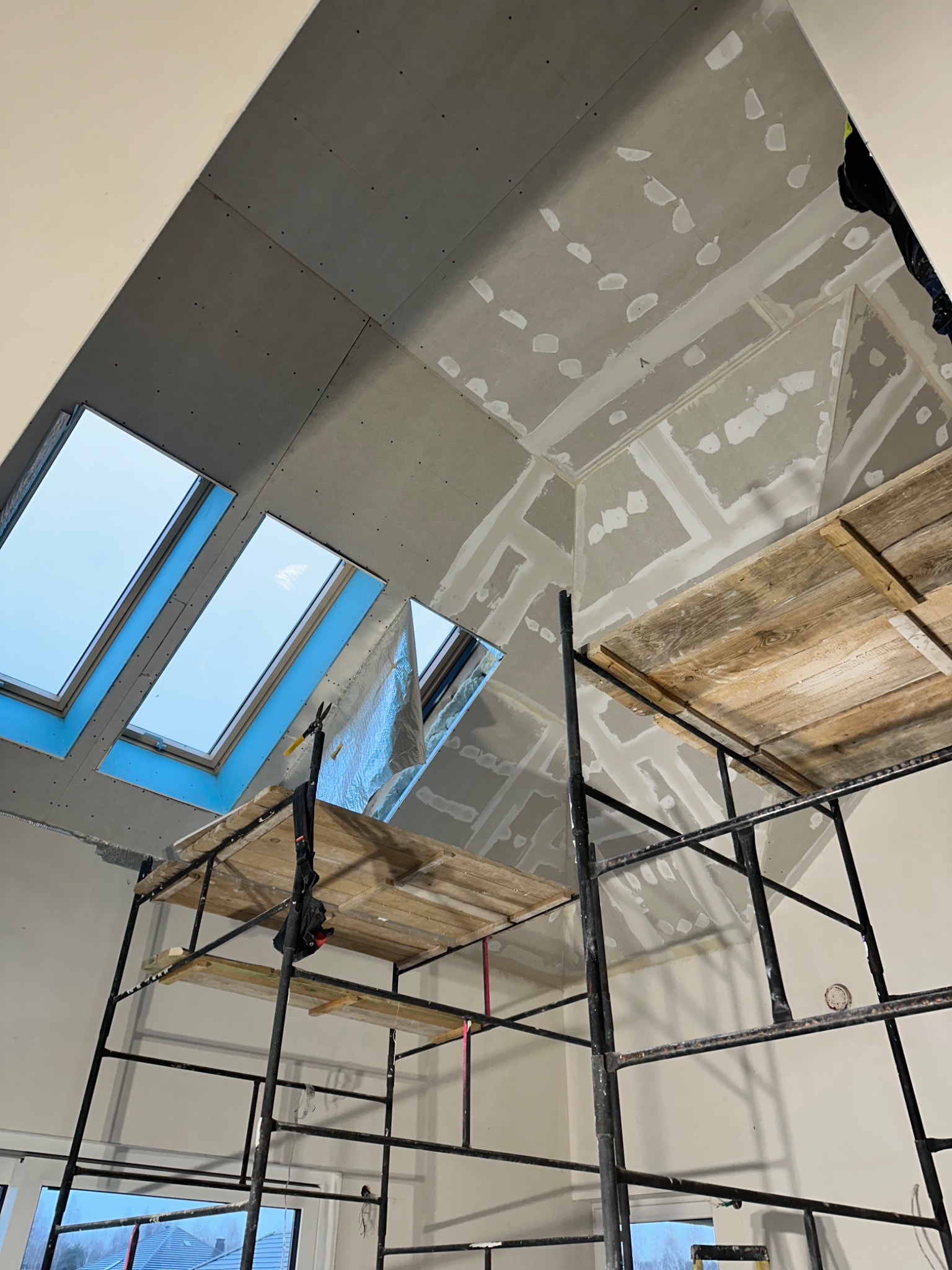 Poddasze z oknami dachowymi w trakcie prac wykończeniowych. Widoczne płyty karton-gips, szpachlowane łączenia i rusztowanie. Ujęcie z dołu, perspektywa wnętrza.