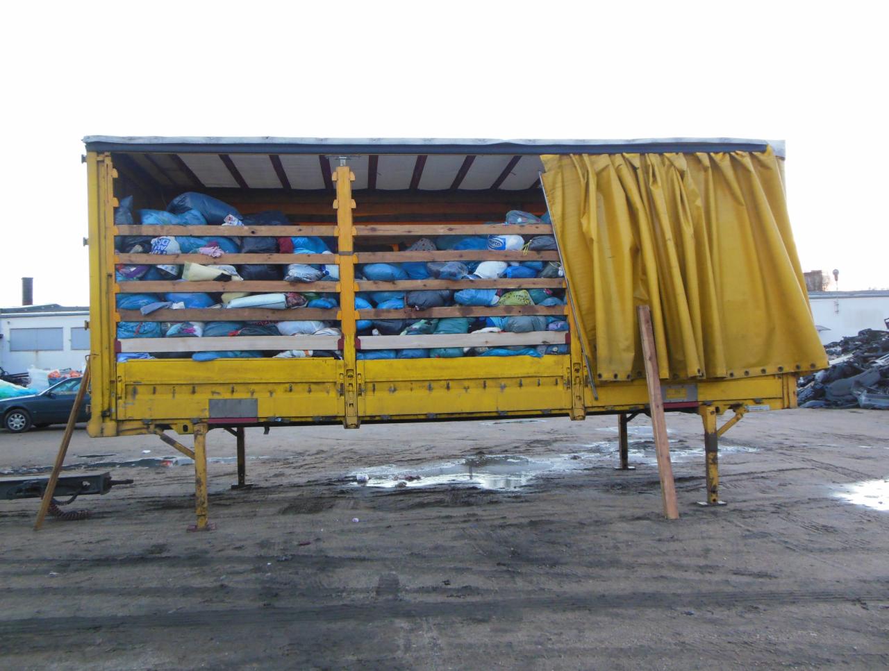 Żółta przyczepa wypełniona workami z tekstyliami, częściowo zasłonięta żółtą plandeką, stojąca na placu z kałużami.