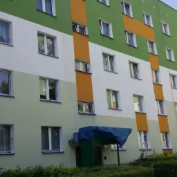 Pracownik w kasku i kamizelce, zawieszony na linach, maluje elewację bloku w odcieniach zieleni, bieli i pomarańczy; widoczne wiadra z farbą i świeżo pomalowany fragment ściany.