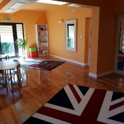 Jasna, pomarańczowa sala z drewnianą podłogą, dywanem z motywem flagi brytyjskiej, stolikami i krzesłami dla dzieci, regałem z książkami i zabawkami, roślinami w doniczkach oraz lustrem w białej...