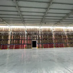 Pełny regał magazynowy z paletami zapakowanych towarów, widok z perspektywy niskiego kąta na posadzkę, oświetlenie górne, metalowa konstrukcja dachu magazynu.