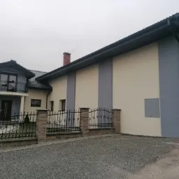 Nowoczesny dom jednorodzinny z dwukolorową elewacją w odcieniach szarości i beżu, widoczny z perspektywy podjazdu z szarego żwiru, z ozdobnym, kutym ogrodzeniem.