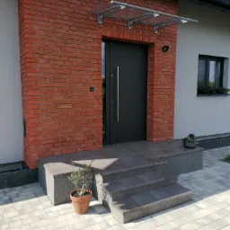 Wejście do domu w Dębowcu z ciemnymi drzwiami, ceglaną wstawką elewacyjną i zadaszeniem, wykończone szarymi płytkami schodowymi i kostką brukową.