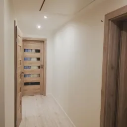 Korytarz z jasnymi ścianami i drewnianymi drzwiami z przeszkleniami, widoczne oświetlenie sufitowe