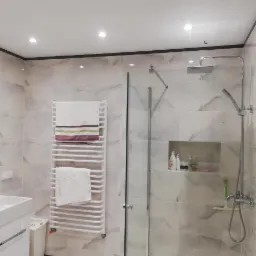 Wykończona łazienka z kabiną prysznicową, umywalką, grzejnikiem drabinkowym z ręcznikami i marmurowymi płytkami ściennymi z ciemną listwą sufitową.