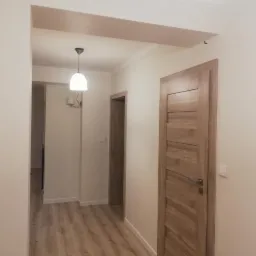 Wykończony korytarz w neutralnych barwach z drewnianą podłogą, drzwiami i listwami. Widoczna lampa sufitowa i wieszak na ubrania w głębi.