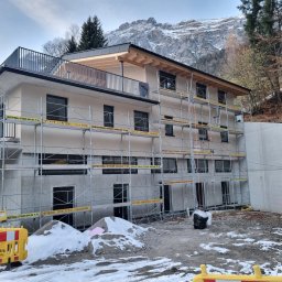 Profi Haus APP Andrzej Paździor - Trzypiętrowy budynek w trakcie budowy, otoczony rusztowaniami z żółtymi tabliczkami, na tle ośnieżonych gór i częściowo ośnieżonego terenu.