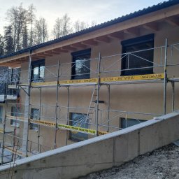 Profi Haus APP Andrzej Paździor - Dom w trakcie ocieplania elewacji, widoczne rusztowania z reklamą firmy, częściowo zakryte okna folią ochronną, pracownik na rusztowaniu.