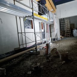 Profi Haus APP Andrzej Paździor - Elewacja budynku w trakcie prac, widoczne rusztowanie z pracownikiem, okno zabezpieczone folią, materiały budowlane na ziemi, drewniana konstrukcja dachu.