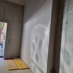 Profi Haus APP Andrzej Paździor - Wnętrze surowego budynku z betonowymi ścianami, jedna ściana pokryta świeżym tynkiem, zabezpieczona żółtymi płytami i czerwoną szczotką, widoczne przewody w folii.