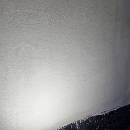 Profi Haus APP Andrzej Paździor - Świeżo wyszpachlowana ściana, oświetlona lampą roboczą, z widoczną folią kubełkową przy podłodze i wystającym przewodem elektrycznym u góry.