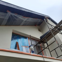 Profi Haus APP Andrzej Paździor - Elewacja domu zabezpieczona folią i taśmą malarską przed natryskowym malowaniem, widoczny pistolet natryskowy, rusztowanie i deski.