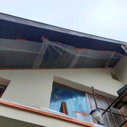 Profi Haus APP Andrzej Paździor - Elewacja domu zabezpieczona folią i taśmą malarską przed malowaniem, widoczne rusztowanie i urządzenie do malowania natryskowego.