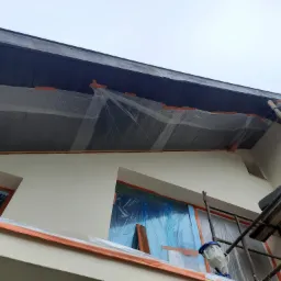 Elewacja domu zabezpieczona folią i taśmą malarską przed malowaniem, widoczne rusztowanie i urządzenie do malowania natryskowego.