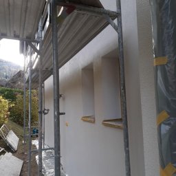 Profi Haus APP Andrzej Paździor - Świeżo otynkowana elewacja domu jednorodzinnego, widok z rusztowania, okna zabezpieczone folią i taśmą malarską, w tle zieleń ogrodu.