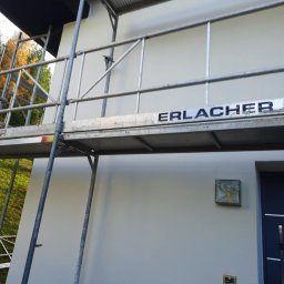 Profi Haus APP Andrzej Paździor - Świeżo otynkowana elewacja domu jednorodzinnego z szarymi detalami, widoczna z poziomu rusztowania firmy Erlacher, z zabezpieczonym oknem i fragmentem ciemnych drzwi.