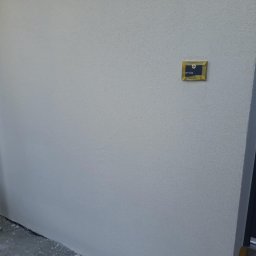 Profi Haus APP Andrzej Paździor - Świeżo otynkowana, jasnoszara elewacja z widoczną fakturą, fragment rusztowania po lewej i ciemne drzwi wejściowe po prawej, mała tabliczka informacyjna na ścianie.