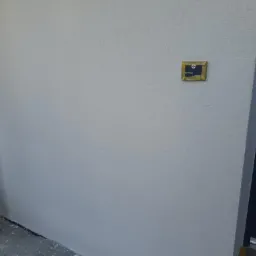 Świeżo otynkowana, jasnoszara elewacja z widoczną fakturą, fragment rusztowania po lewej i ciemne drzwi wejściowe po prawej, mała tabliczka informacyjna na ścianie.