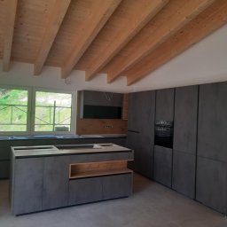 Profi Haus APP Andrzej Paździor - Wnętrze kuchni w trakcie montażu: szare, matowe szafki kuchenne z drewnianymi akcentami, wyspa kuchenna bez blatu, widoczne okno z zielenią na zewnątrz, surowy drewniany strop z belek.