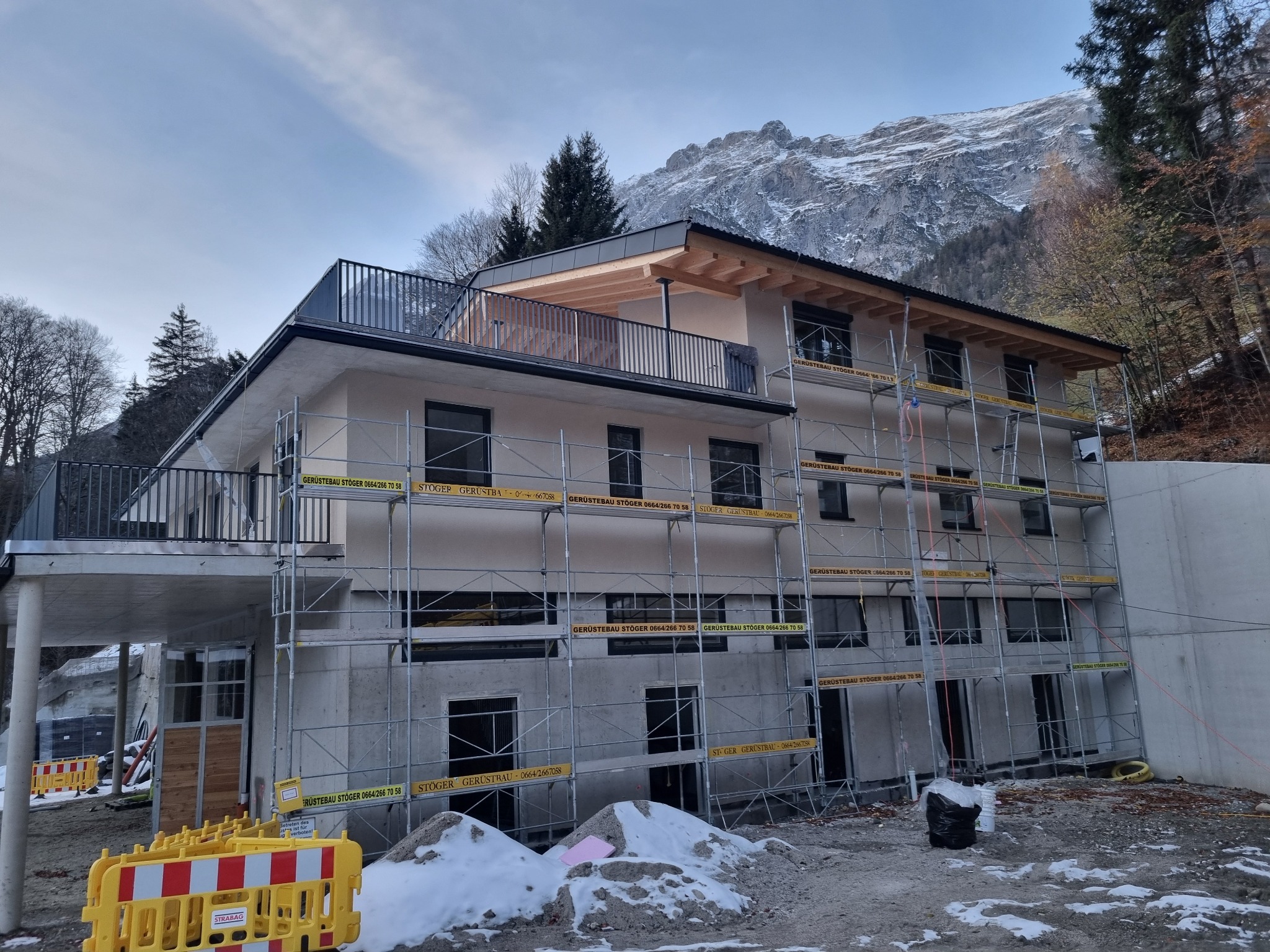 Nowoczesny, dwupiętrowy dom w trakcie budowy, z widocznymi rusztowaniami i częściowo wykończoną elewacją na tle górskiego krajobrazu.