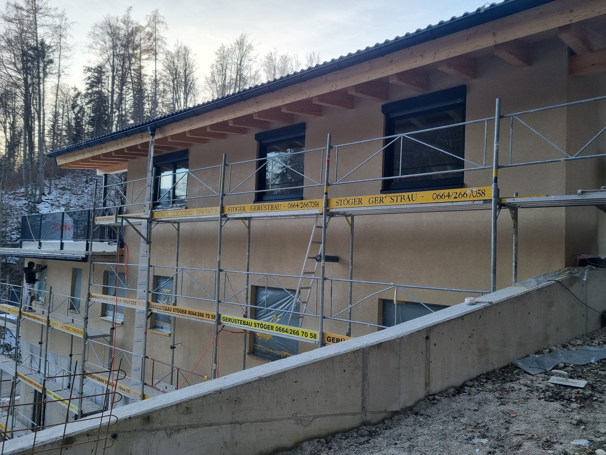 Dom w trakcie ocieplania elewacji, widoczne rusztowania z reklamą firmy, częściowo zakryte okna folią ochronną, pracownik na rusztowaniu.