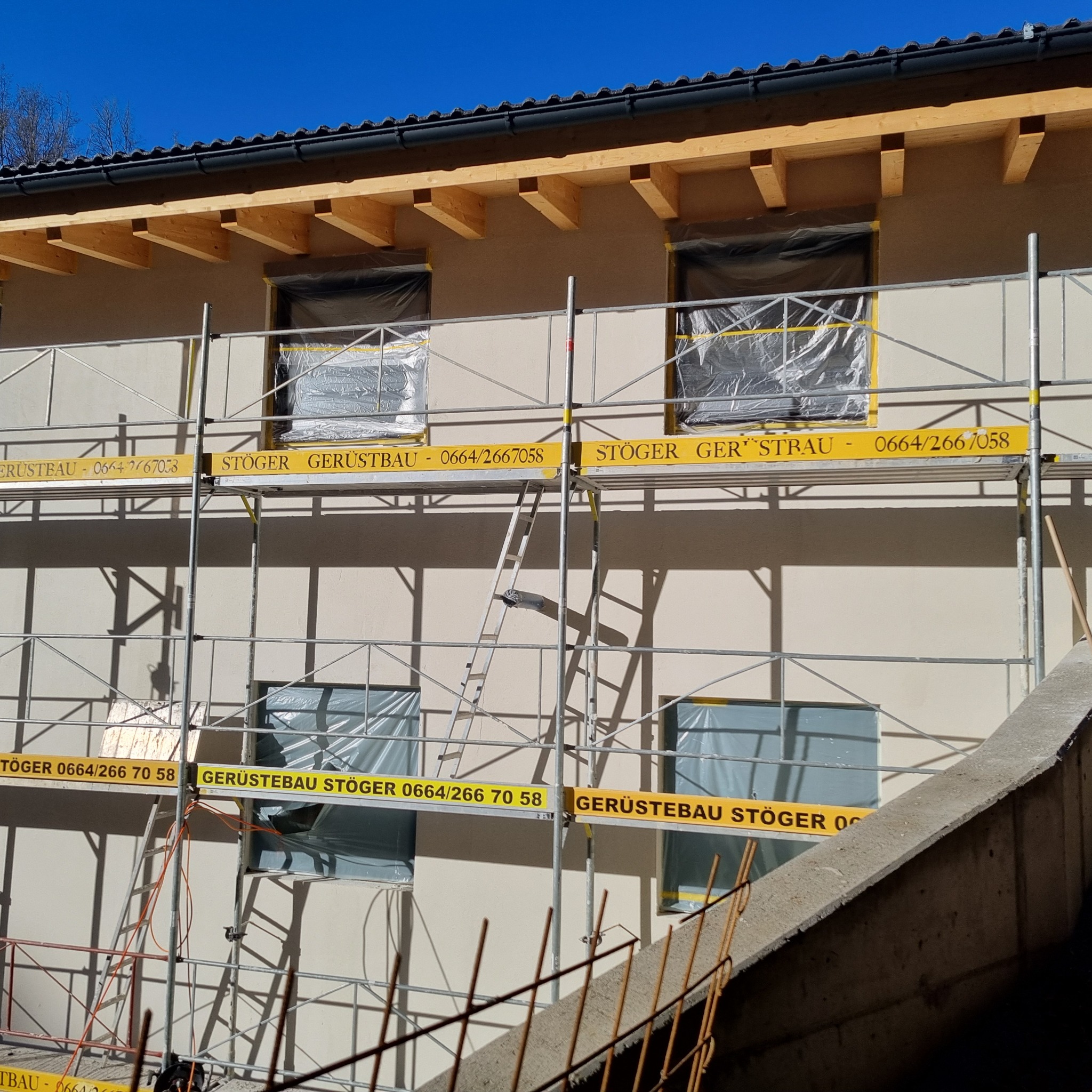 Elewacja budynku w trakcie przygotowań do prac, okna zabezpieczone folią, widoczne rusztowania z żółtymi tablicami reklamowymi firmy Gerüstbau Stöger, fragment betonowej konstrukcji w prawym dolnym...