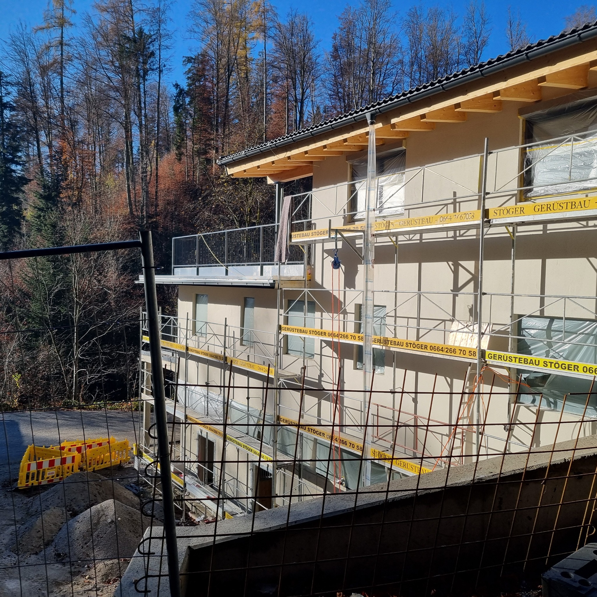 Trzy kondygnacyjny dom w trakcie prac elewacyjnych, osłonięte okna folią, rusztowania z widocznym brandingiem firmy, w tle las i barierki ostrzegawcze.